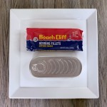A closed can of Beach Cliff herring fillets and their outer wrapper on a square white plate.